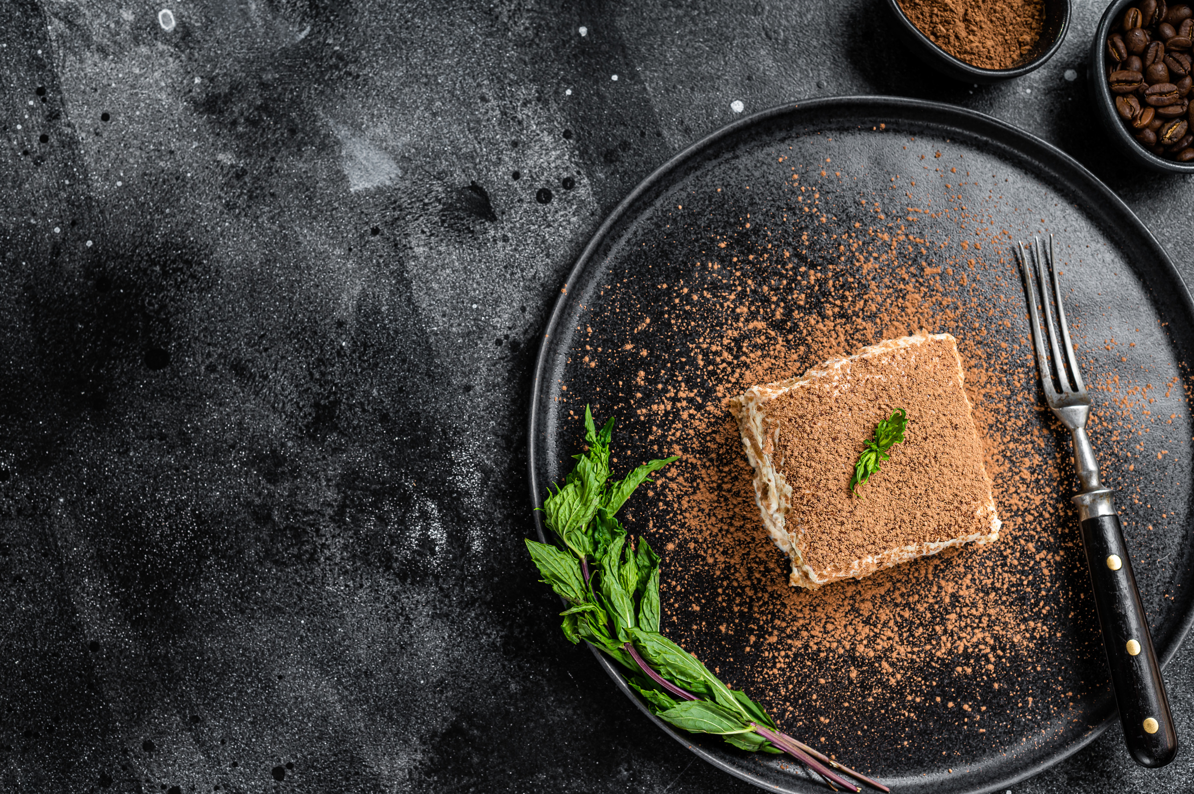 Italian Tiramisu cake with cocoa and mint on a plate. Black background. Top view. Copy space